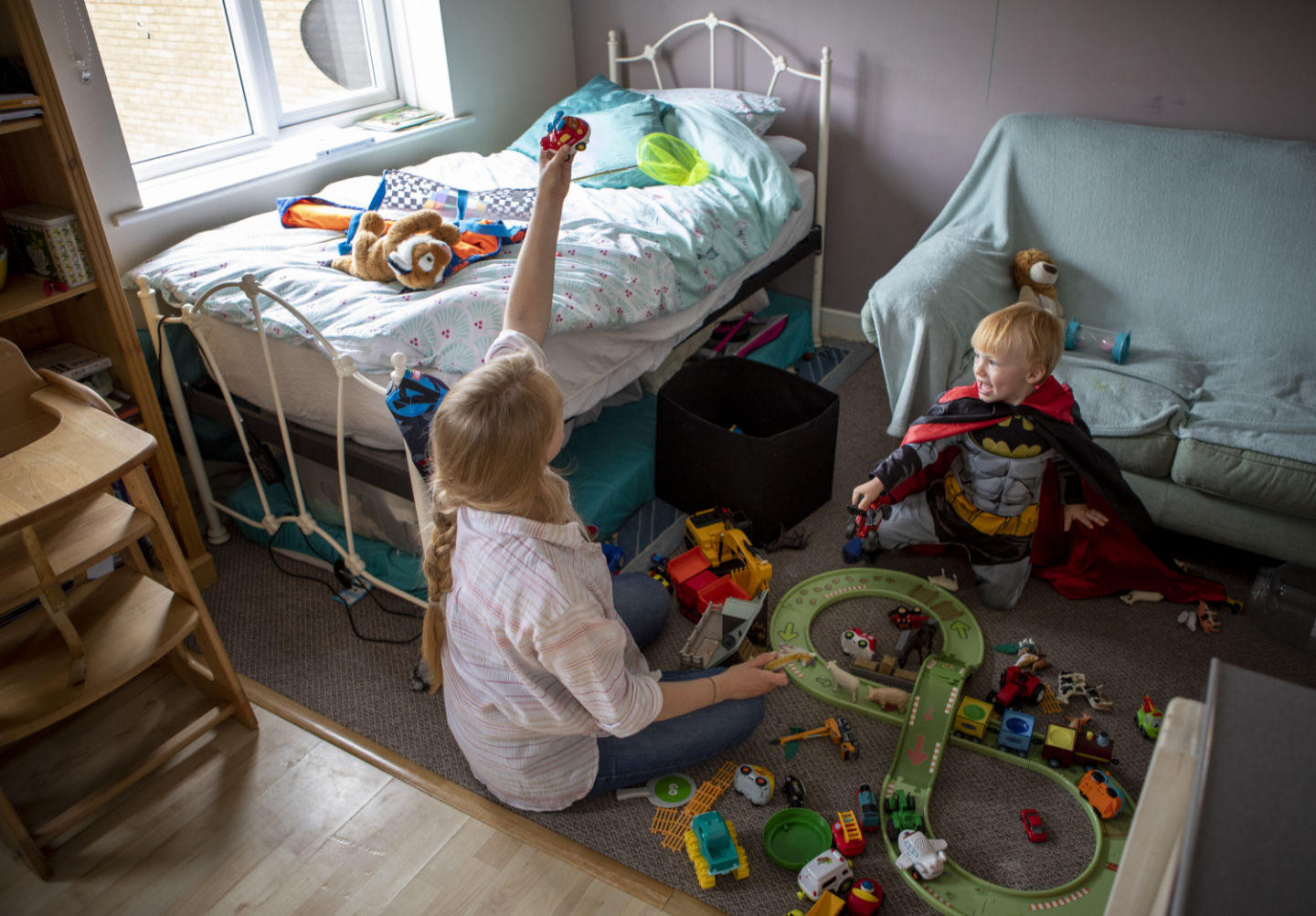 A mother and her little boy, dressed as a superhero, play with his toys in a small room