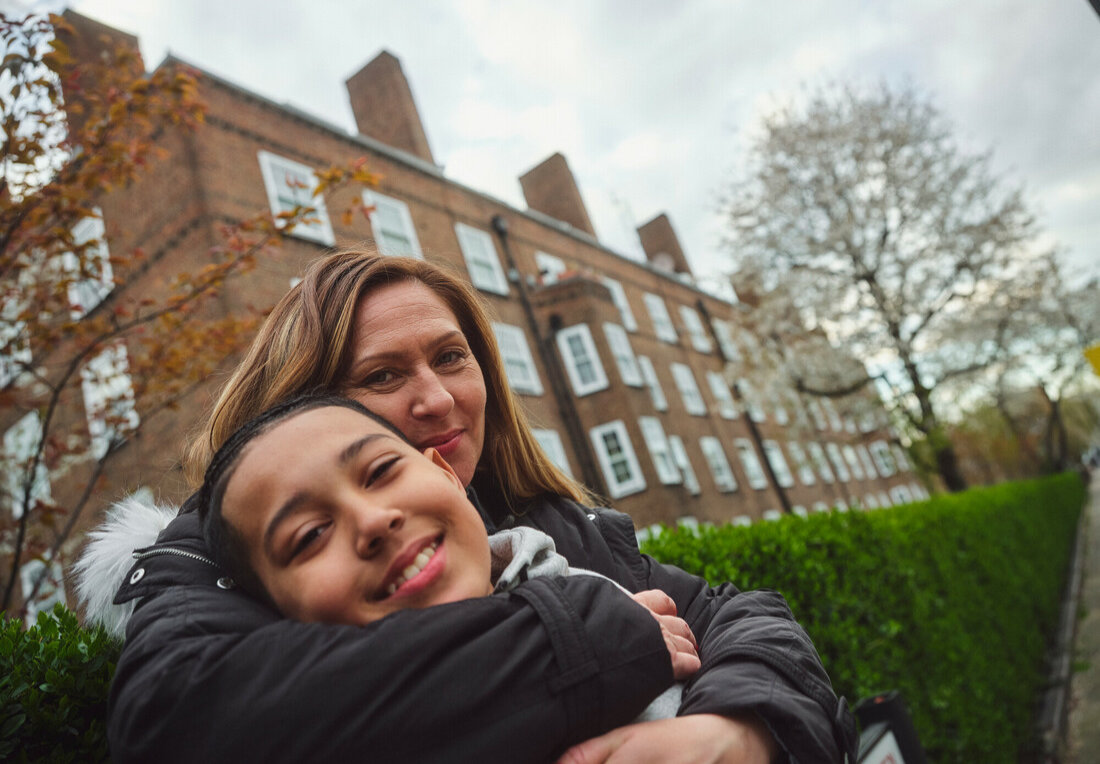 Mother hugging daughter, both looking at the camera, standing outdoors in front of a housing complex.