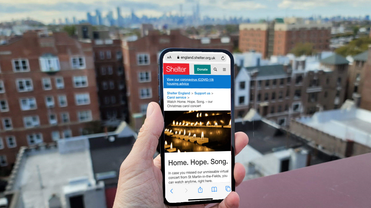 A hand holds a mobile phone showing a page of a Shelter event, overlooking buildings.