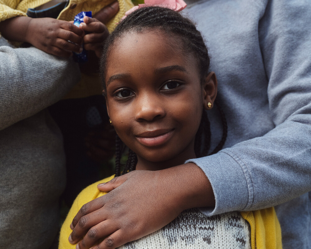 Young girl looking at camera, with her mother's arm holding her.