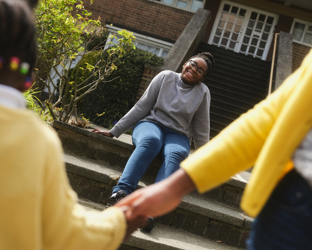 A mother sitting on steps outside a housing complex. In the foreground are the arms of her two young daughters.