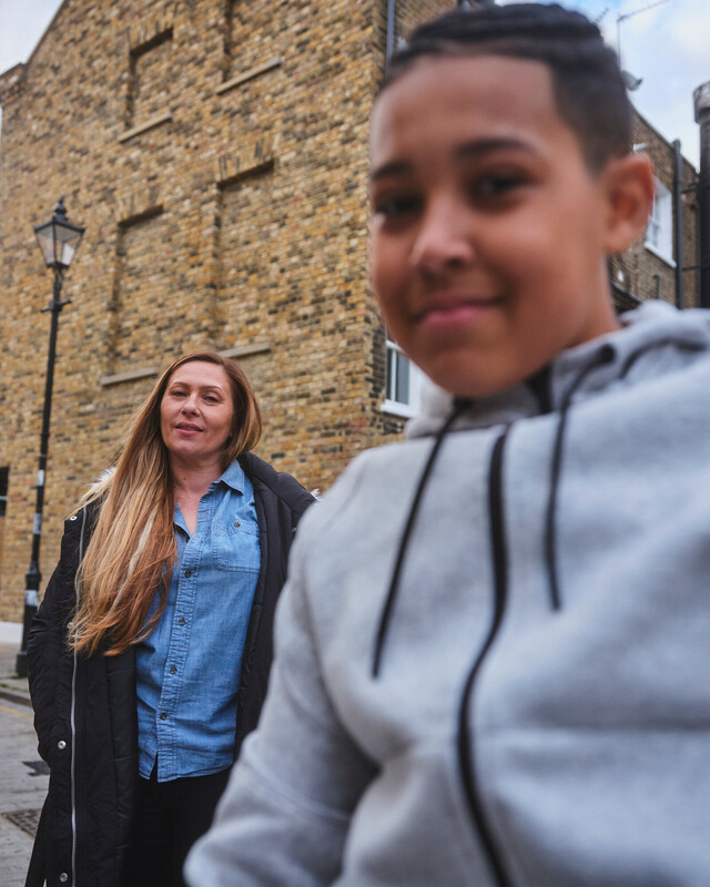 Outdoor photo of a boy in the foreground and his mum in the background. Behind them is a brick building.