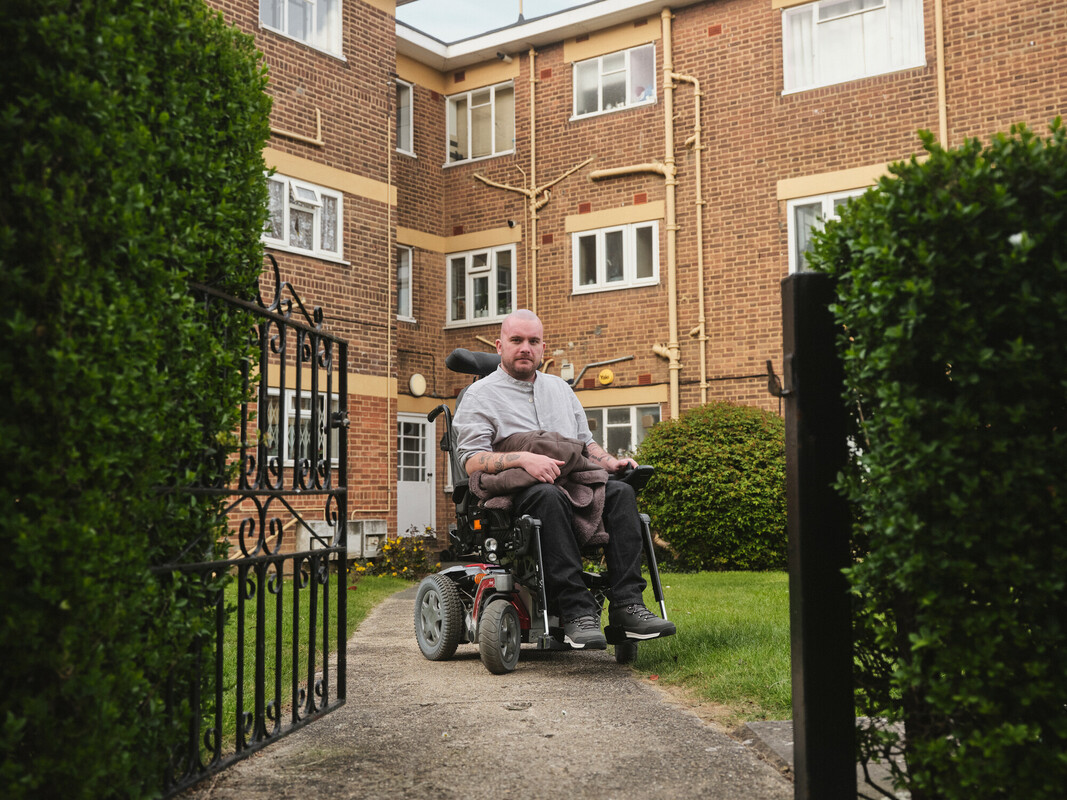 Man in a wheelchair inside the gate of a 3-story housing complex