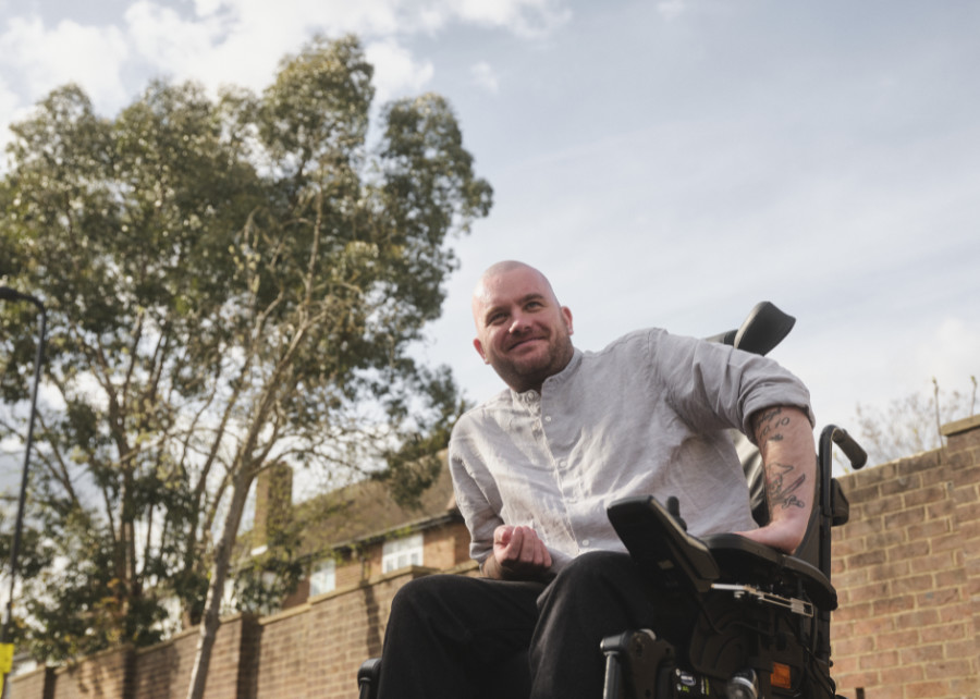Man in a wheelchair, outdoors with a brick wall and trees behind him.