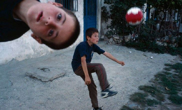 Photo by Alex Webb of a boy on a gravel forecourt kicking a football. In the foreground, appearing from side of phot is another boy.