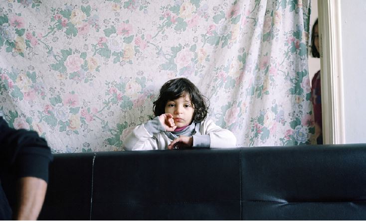 Photo by Edward Thompson of a small boy indoors, looking at camera in front of a floral pattern curtain. The arm of a man is in the foreground, and a woman peers from behind the curtain.