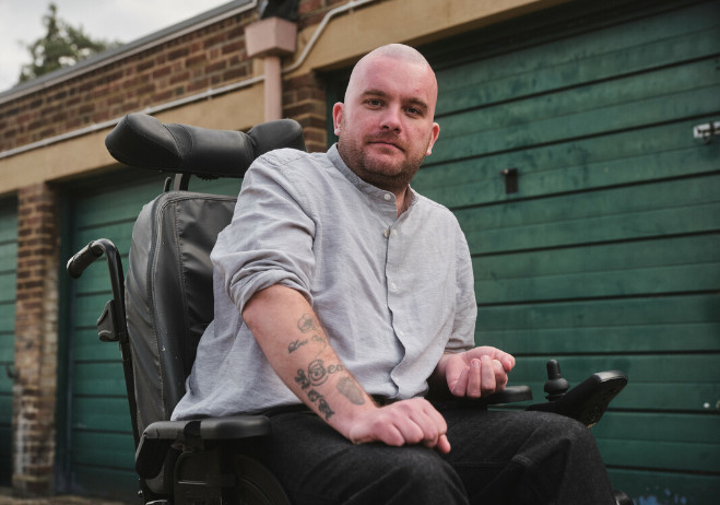 Outdoor shot of a man in a wheelchair looking at the camera. Behind him is garage door.