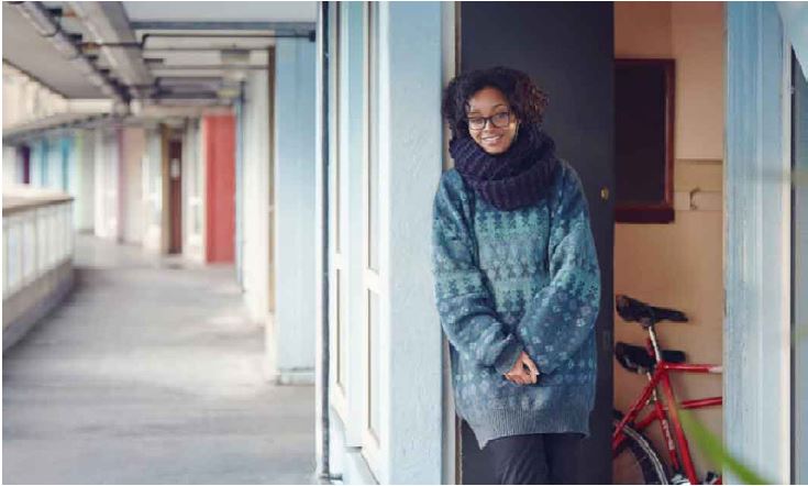 Photo by Tom Hull of a woman in a blue jumper, blue scarf and glasses, standing outside by her flat's door, smiling and facing camera,