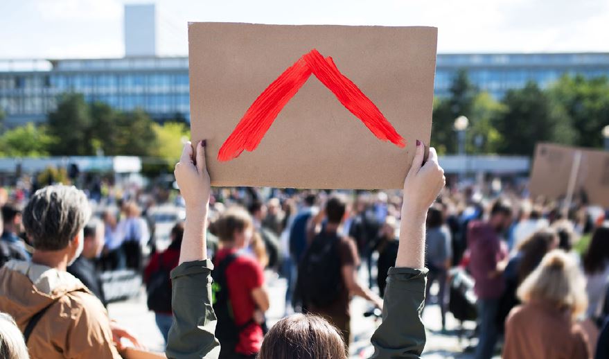 Photo of a person in a crowd holding up the Shelter symbol on a cardboard sign